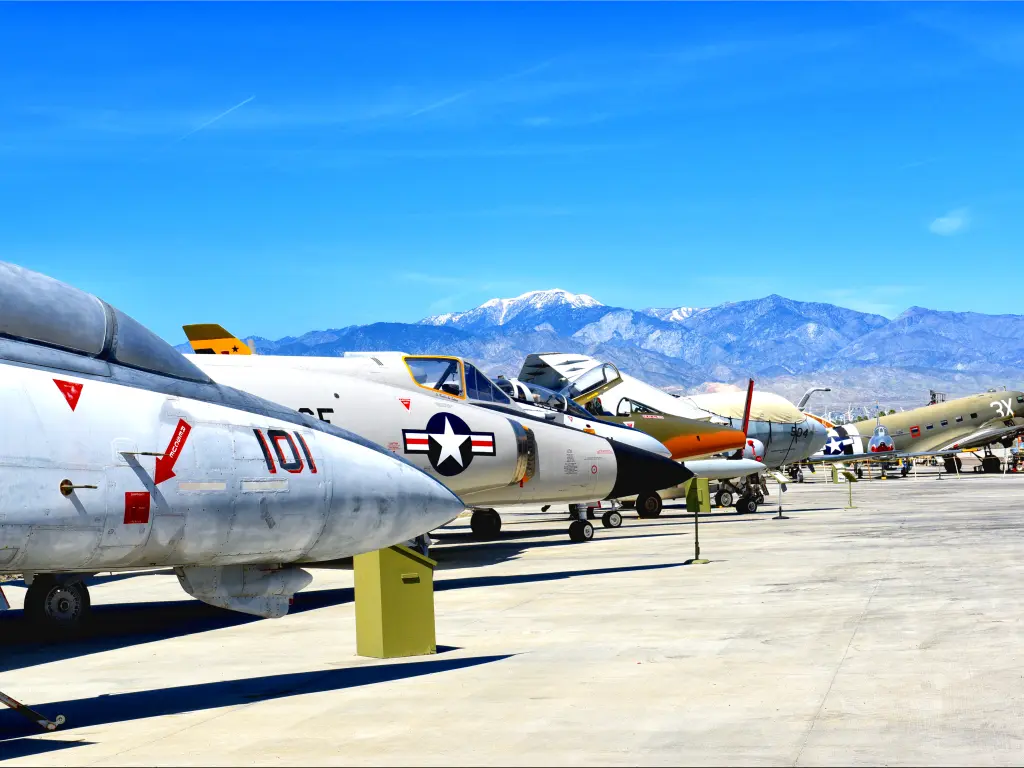 A line of vintage flyable WWII aircraft displays outside on a sunny day with a view of the mountains from afar at Palm Springs Air Museum.