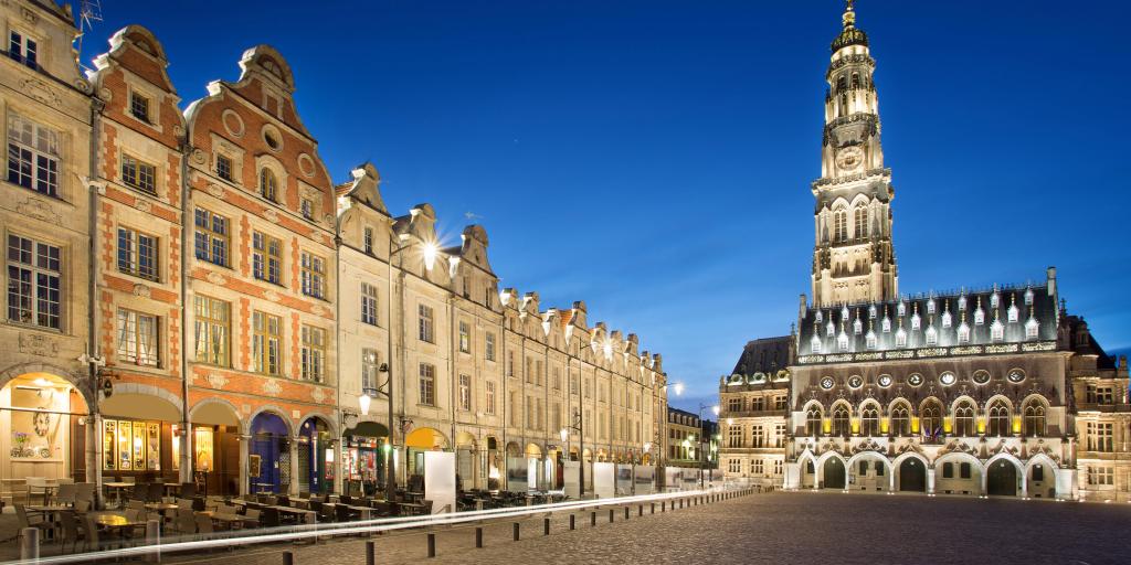 A row of townhouses with curved roofs on Place des Heros, Arras, at night, with the rebuilt belfry in the background. 