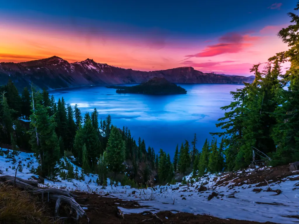 Crater Lake in winter at sunset, with a pink-hued sky and snow on the ground between the pine trees