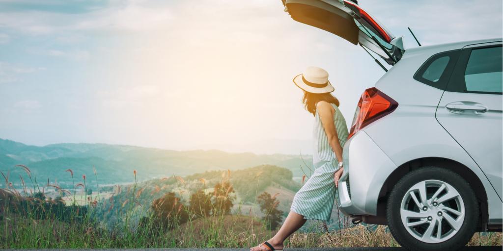 Woman traveller sitting on hatchback car with mountain scenery in the background 