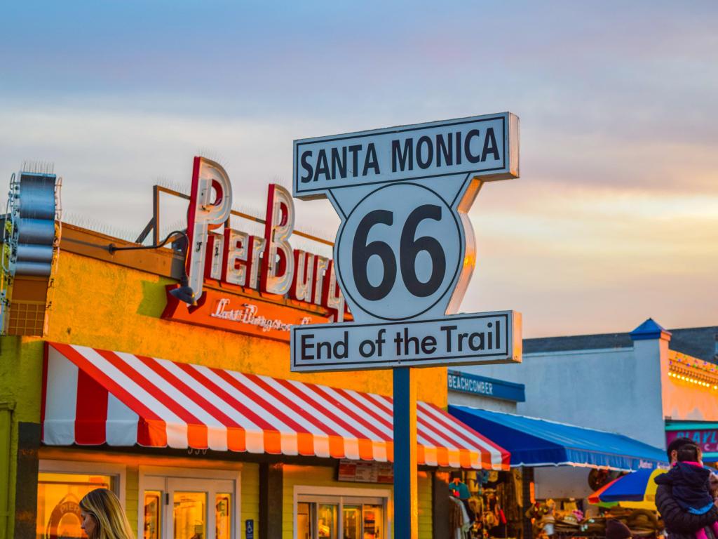 Route 66 End Sign on Santa Monica Pier, California, with brightly colored food stands in the background on a sunny day