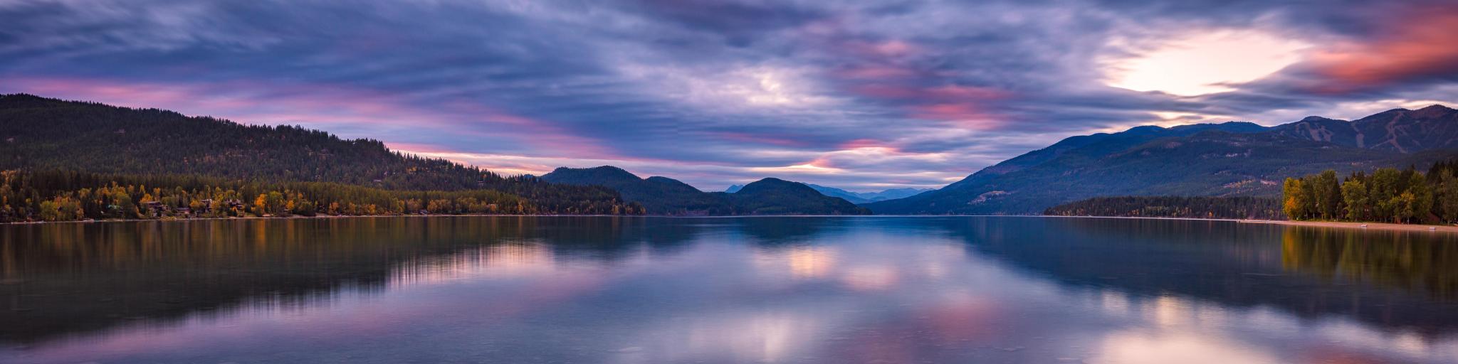 Whitefish Lake, Montana with a stunning sunset sky of pinks and purples, mountains in the distance reflecting in the water and green land either side.