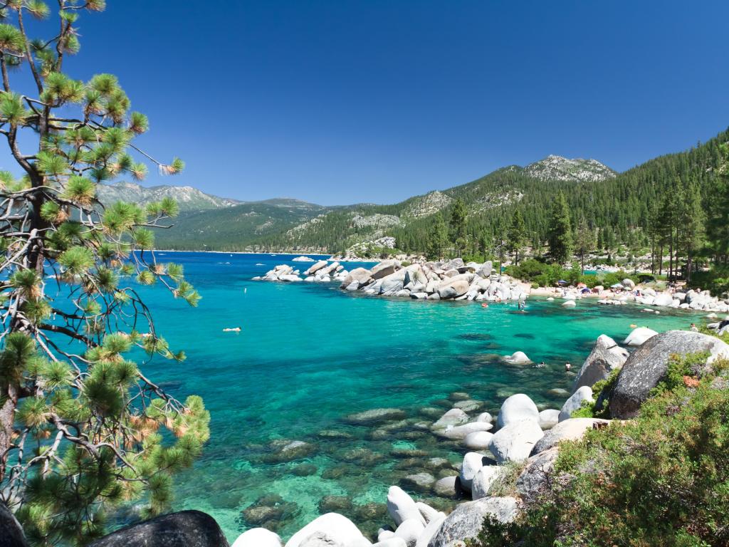 Sand Harbor, Lake Tahoe, USA with rocks and trees in the foreground, a stunning turquoise sea and mountains in the distance covered in trees on a clear sunny day.