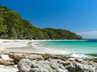 Jervis Bay, Australia with a stunning beach surrounded by rocks in the foreground, crystal clear water and dense woodland in the background.