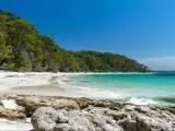 Jervis Bay, Australia with a stunning beach surrounded by rocks in the foreground, crystal clear water and dense woodland in the background.