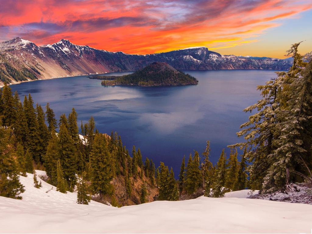 Crater Lake at sunset, with a pinkish light cast over the snow that covers the mountains and pine trees