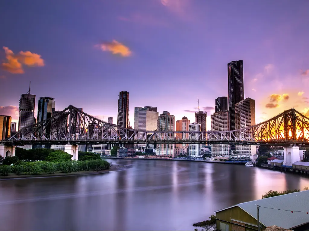 Bridge across steel grey river with tall buildings on the bank, purple blue sky and bright light of setting sun