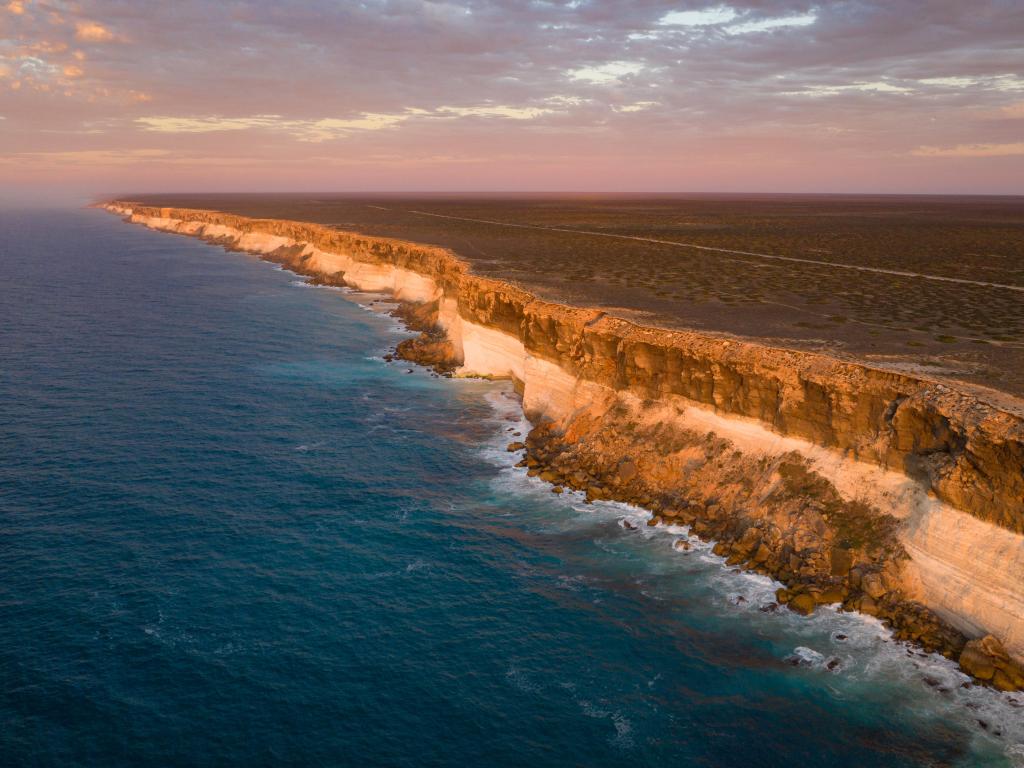 Tall cliffs with lines of different coloured rocks, with ocean breaking at the bottom and wide, flat, dry ground at the top