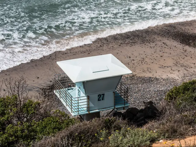 An aerial shot of a pale blue and white wooden lifeguard tower stands on the sands at South Carlsbad State Beach, with vegetation growing behind it