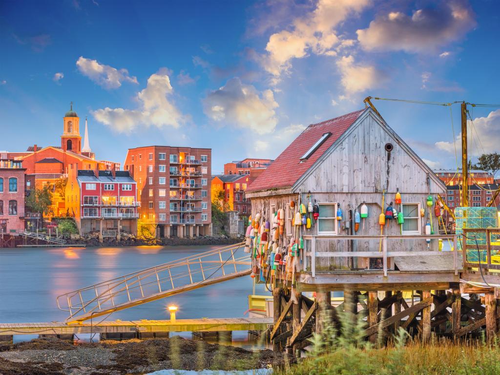 Wooden boat house and pier adorned with colourful buoys, beside the river with low rise buildings on the far bank