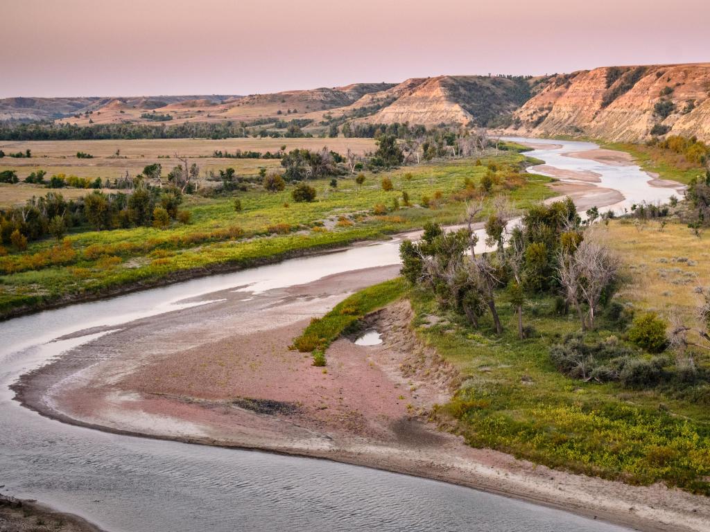 Theodore Roosevelt National Park, North Dakota with a river running through and flat green land at either side leading to mountains in the distance at sunset.