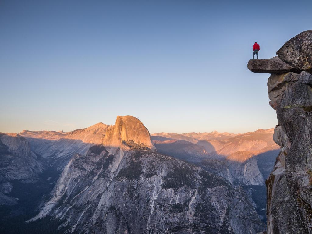 A fearless male hiker is standing on an overhanging rock at Glacier Point enjoying the view over Half Dome at sunset 