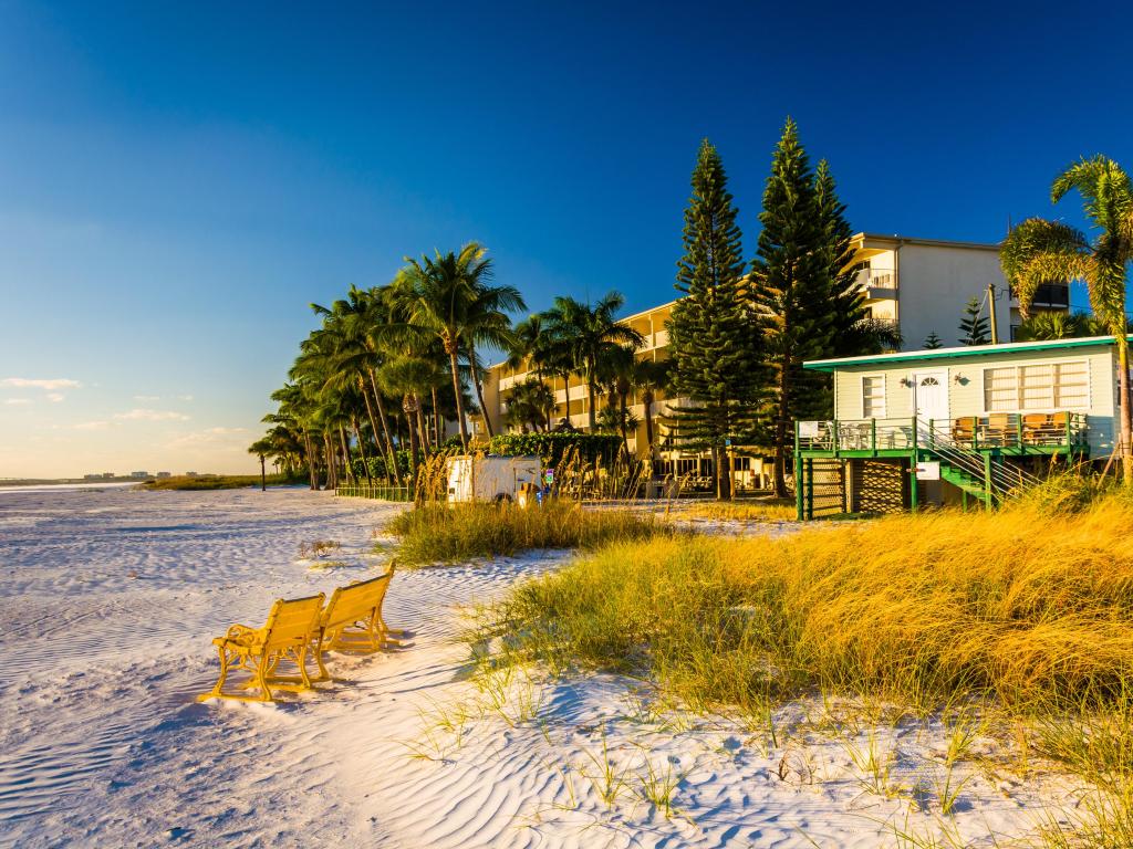 Fort Myers Beach, Florida with sand dunes, chairs facing the ocean and beach buildings behind a row of palm trees. 