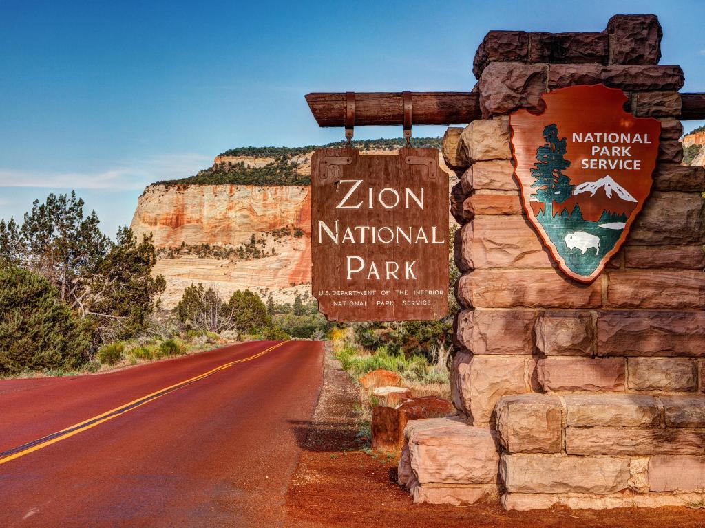 East Entrance Zion National Park Sign in the forefront, with the towering red rocks in the background shot, Utah