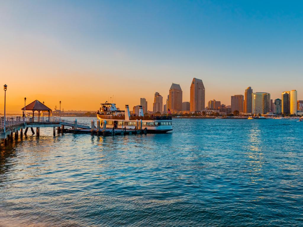 High rise buildings lit up with orange sunset light, viewed across the deep blue slightly rough ocean with a ferry docked at a wooden pier