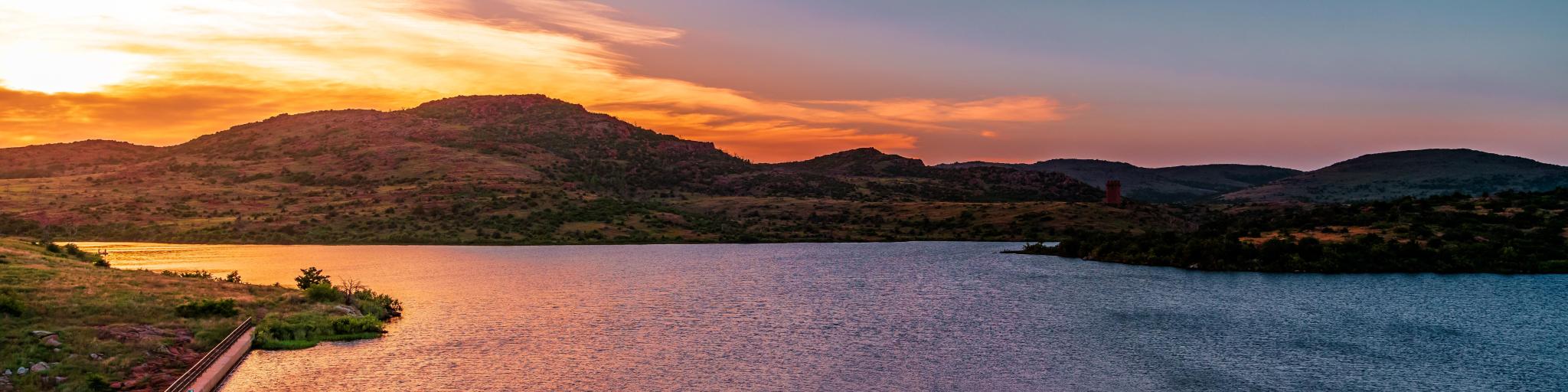 Wichita Mountain Wildlife Preserve, Oklahoma, United States with rock formations in the foreground, a large lake and mountains in the distance at sunset.