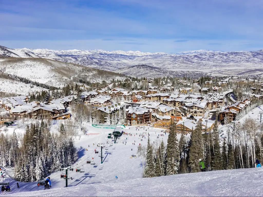 Deer Valley Ski Resort, Utah with a chair lift, ski slopes in the foreground and the ski resort before the snow covered mountains in the distance on a sunny day.