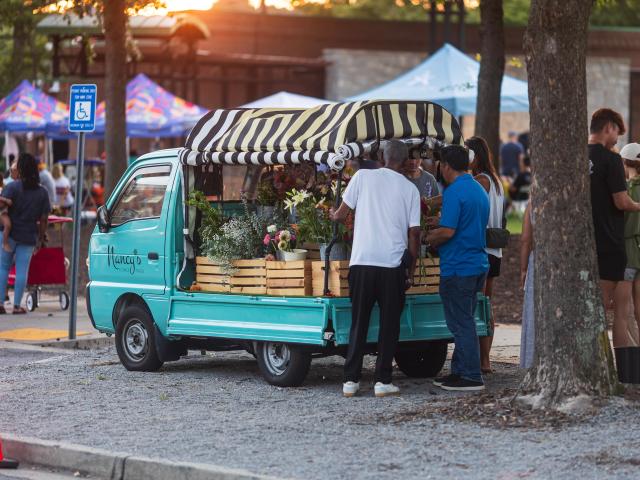 Customers buying flowers from a blue-colored truck during a festival with sun setting in the background