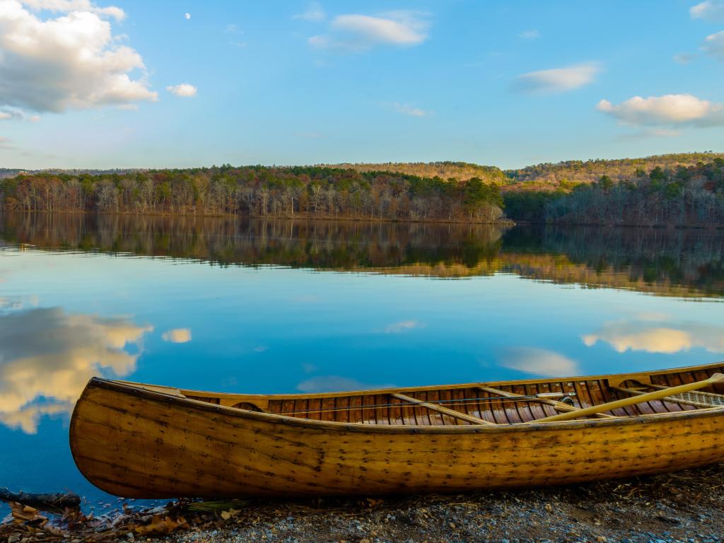Oak Mountain State Park, Birmingham, US at autumn with a wooden boat in the foreground on land, beautiful water reflecting the blue sky and trees in the background.