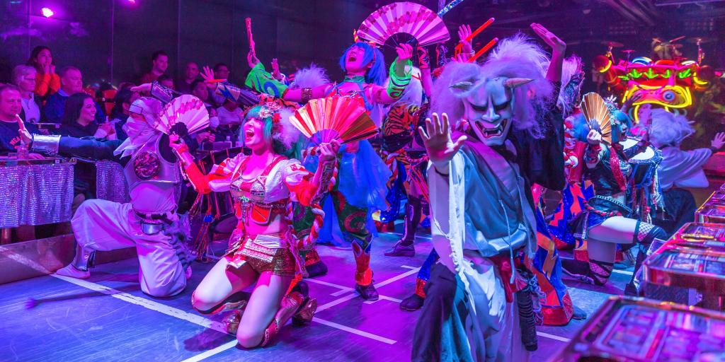 Actors at the robot restaurant in Tokyo dancing - in the front is a man in a Halloween mask with horns and a girl with blue hair holding fans