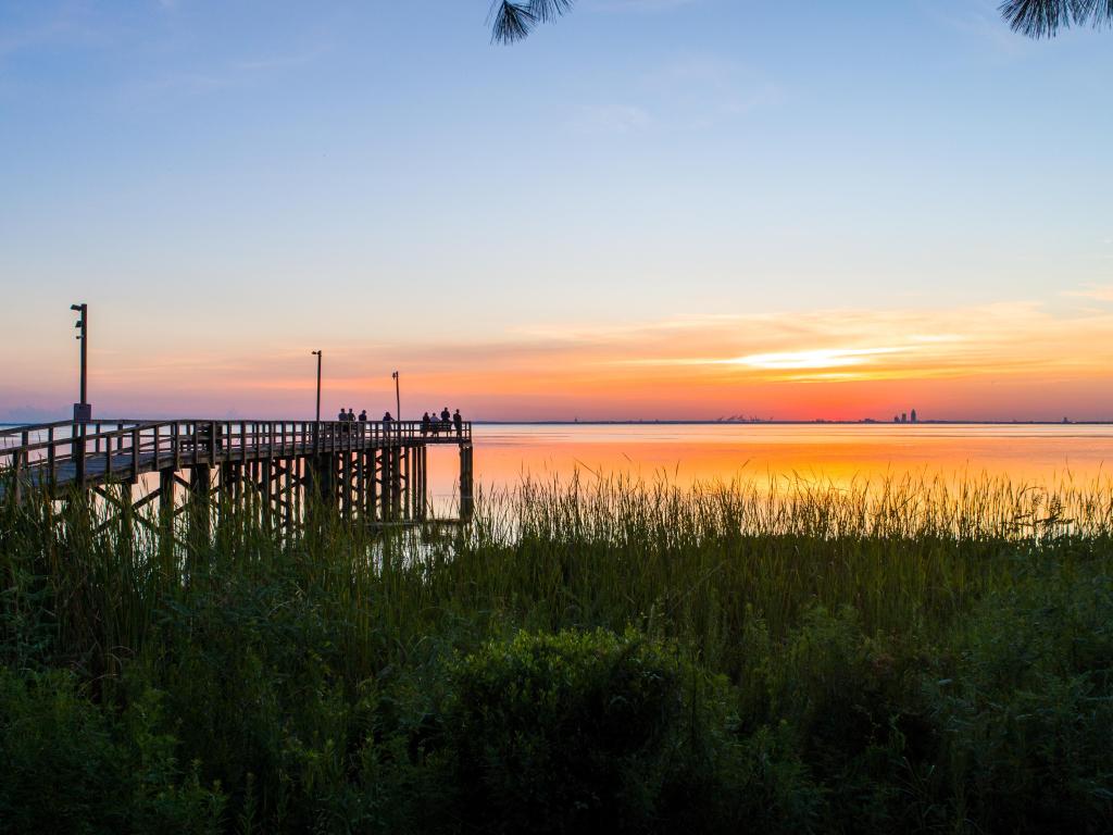 Wooden pier and grassy shore silhouetted against wide view of gold and pink water across Bay