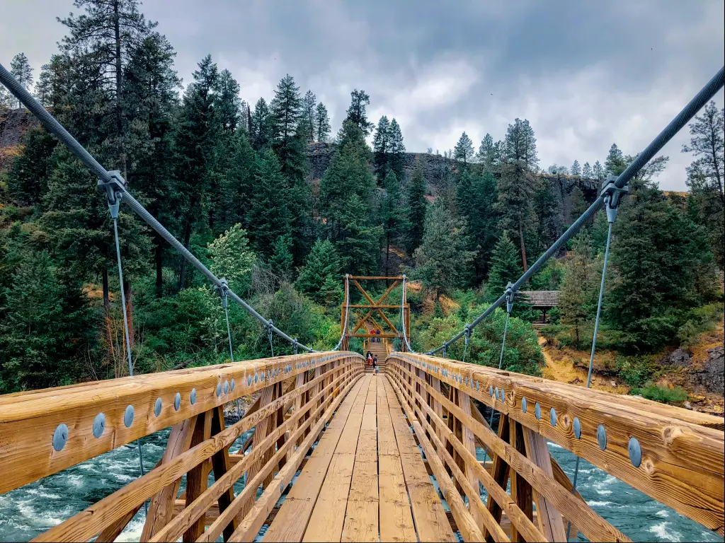 Two people on the other side of the wooden swinging bridge with tall green trees in their back and raging water in Riverside State Park, Spokane, Washington