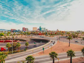 A scenic view of the cars driving along the intersecting highway with palm trees and bushes planted along the road with a picture of the skyscrapers of Phoenix in a fine day with a thin sheet of clouds covering the blue sky