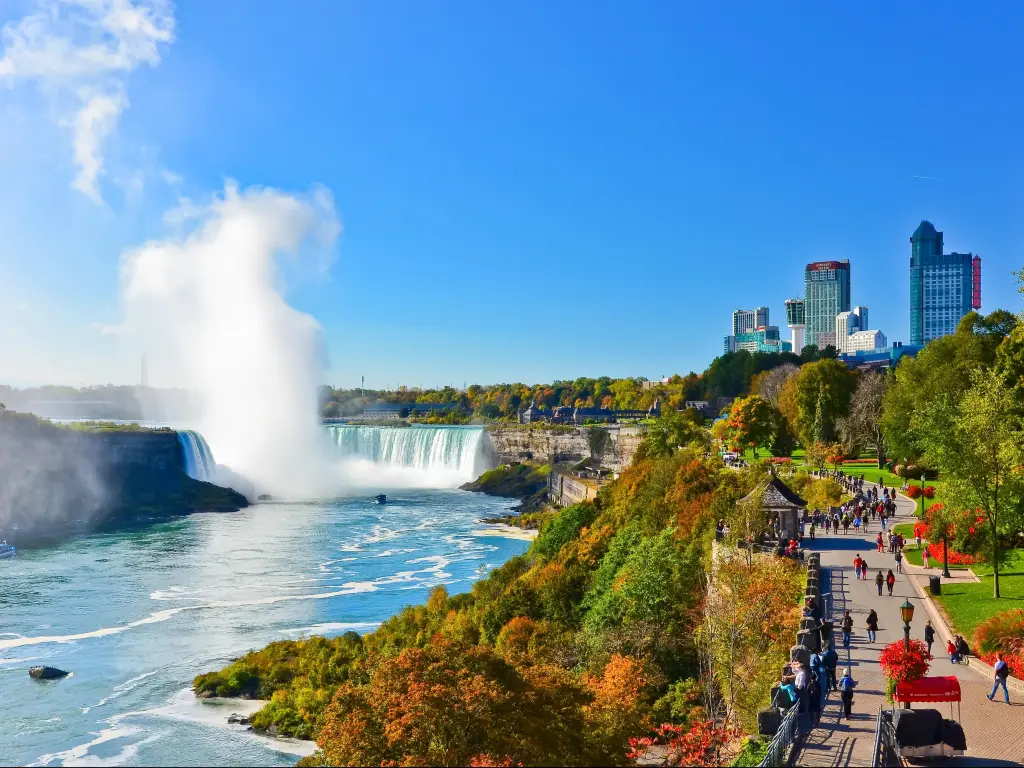 Niagara Falls, Canada with a view of Niagara Falls on a sunny day in fall in Canada with the buildings in the background and a path with people to the edge.