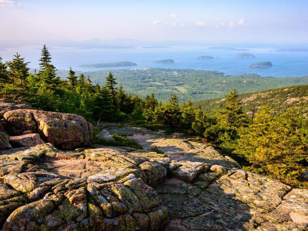 Acadia National Park, USA with large rocks in the foreground, dense forests and lakes beyond taken on a sunny day.
