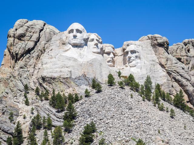 Shows the faces of past US presidents carved on granite as a massive sculpture on Mount Rushmore during a sunny day