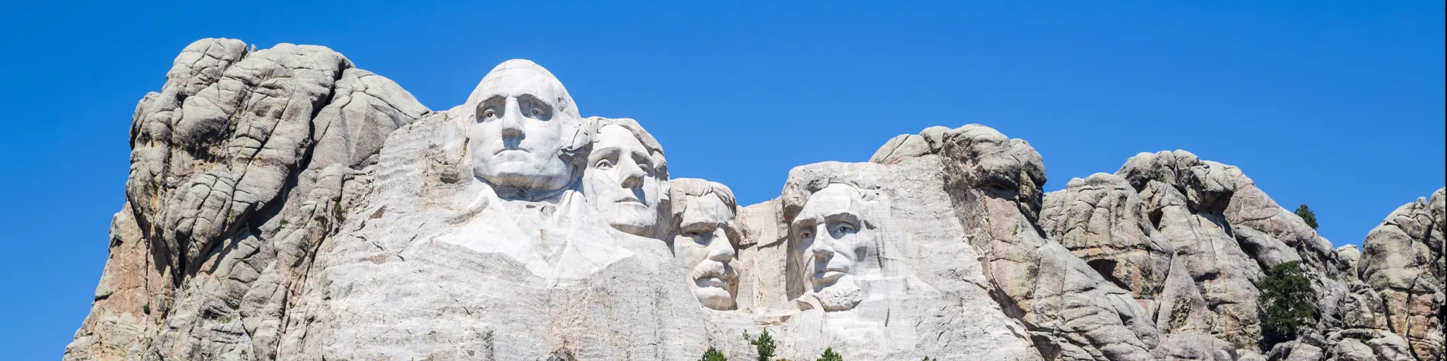Shows the faces of past US presidents carved on granite as a massive sculpture on Mount Rushmore during a sunny day