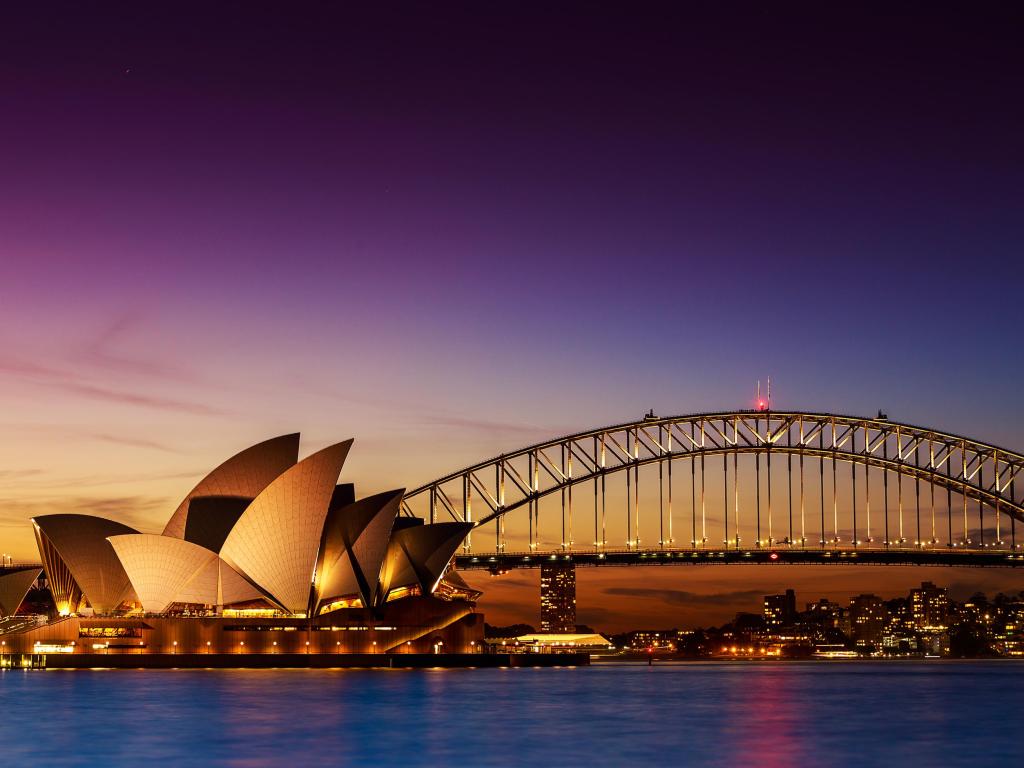 Sail-shaped outline of Sydney Opera House next to Harbour Bridge illuminated in front of dark night sky and high rise buildings in the background with light