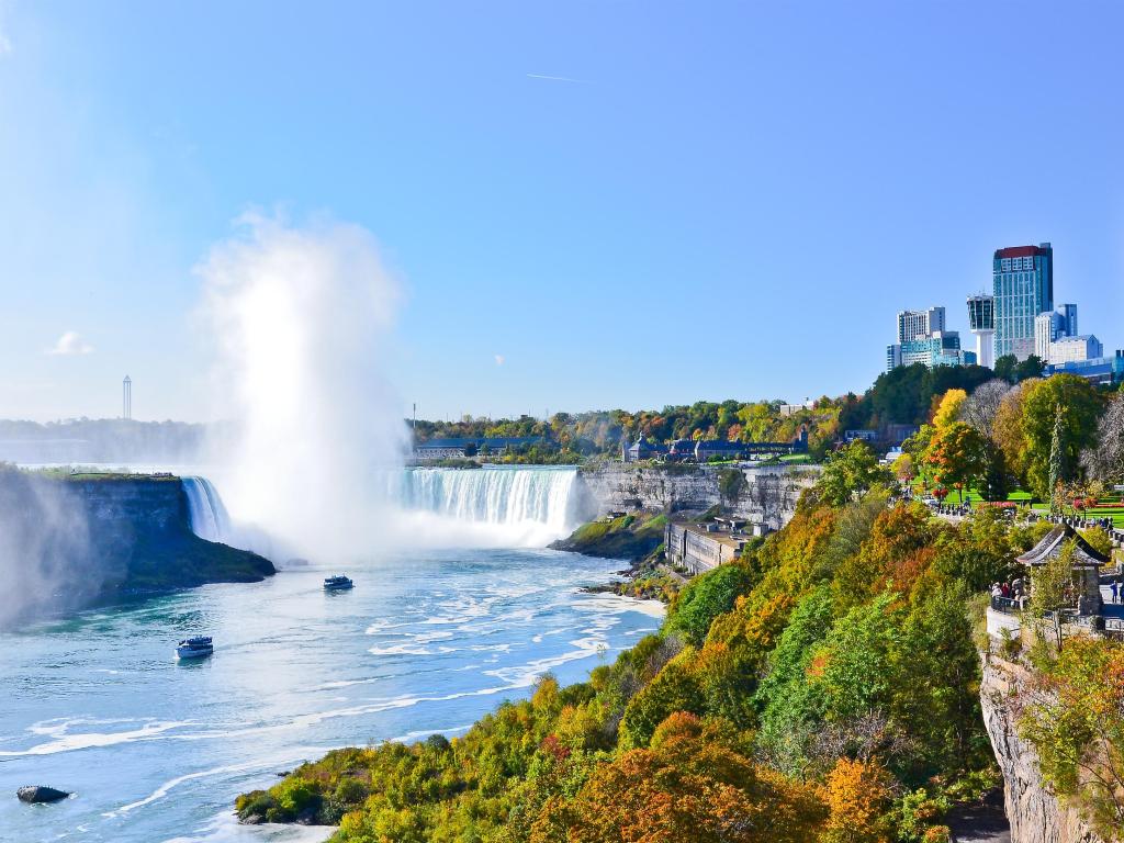 Niagara Falls, USA/Canada with a view of the Falls in fall, golden trees in the foreground and water spray in the distance, taken on a clear sunny day.