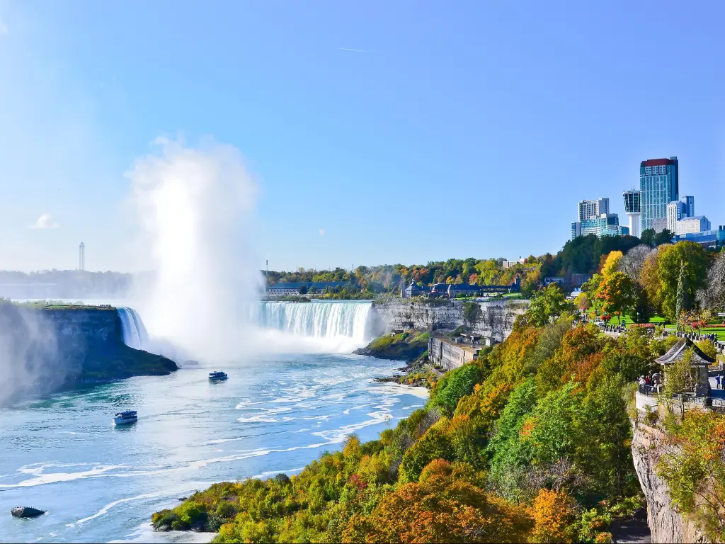 Niagara Falls, USA/Canada with a view of the Falls in fall, golden trees in the foreground and water spray in the distance, taken on a clear sunny day.