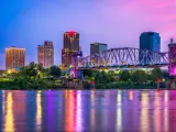 Little Rock, Arkansas, USA downtown skyline on the Arkansas River at night with the buildings lit up in blues, pinks and reds and reflecting in the water.