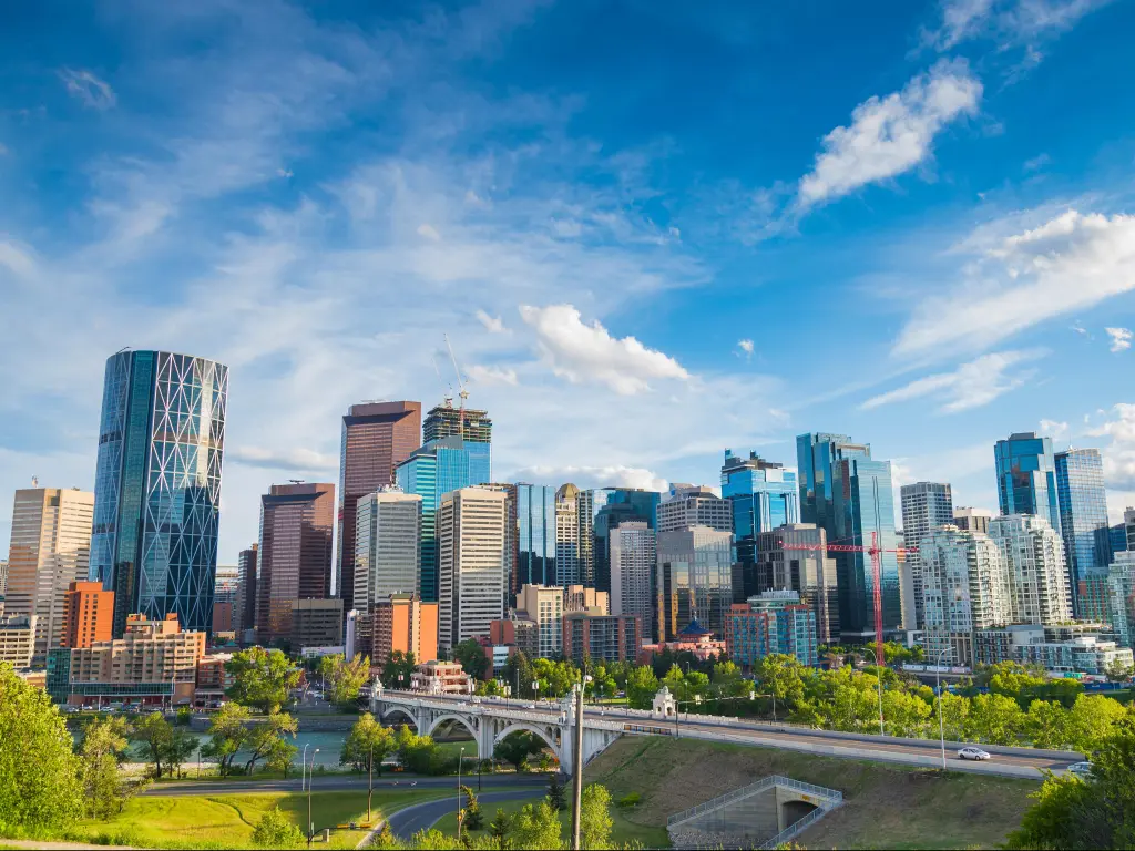Calgary, Alberta, Canada with the city skyline in the background against a cloudy but blue sky, green grass and a road bridge in the foreground. 