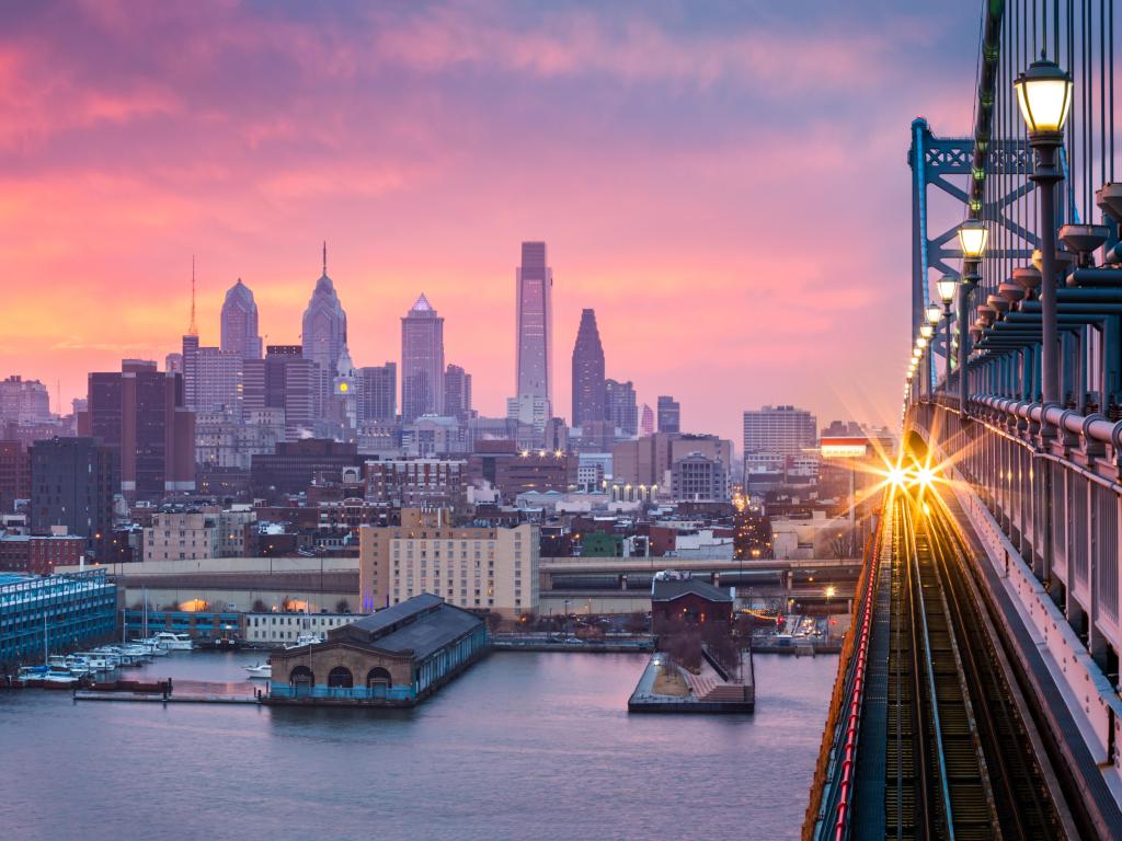 Philadelphia, USA with a panoramic view of the city skyline in the distance under a hazy purple sunset with an incoming train crossing the Ben Franklin Bridge.