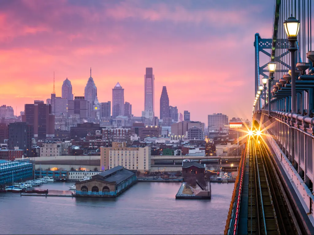 Philadelphia, USA with a panoramic view of the city skyline in the distance under a hazy purple sunset with an incoming train crossing the Ben Franklin Bridge.