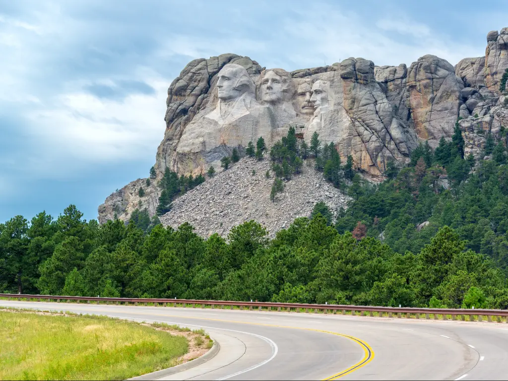 An empty curve road leading to Mount Rushmore National Monument in South Dakota and a view of the carved faces of the Four US Presidents in the mountain and green trees