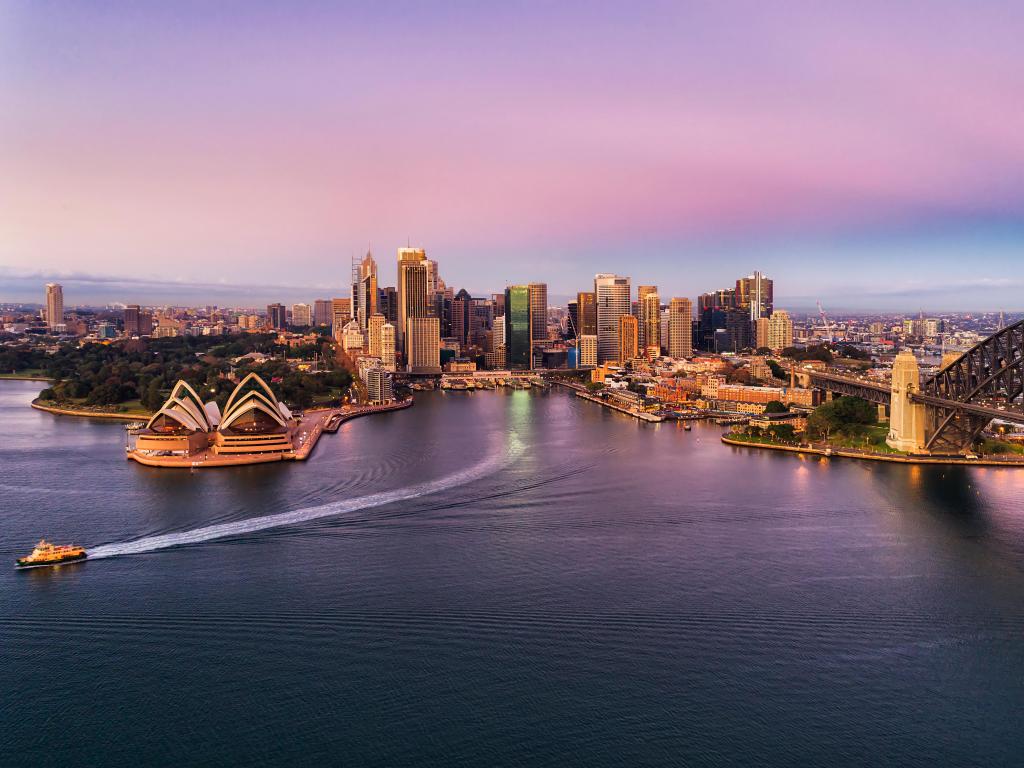 Sydney Harbour, Australia  taken with a pink colourful sunrise over Sydney city CBD landmarks on Sydney Harbour shores touched by steel arch of the Harbour bridge with passenger ferry on route to wharf.