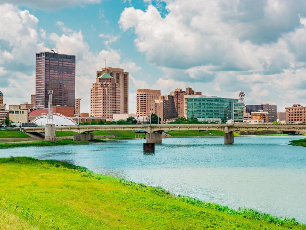 Dayton, Ohio with the Miami River in the foreground and a bridge with the city buildings in the background against a blue sky. 