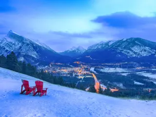 Two red chairs on a hill in the snow, overlooking the city as the night falls
