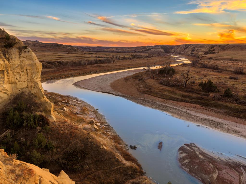 Theodore Roosevelt National Park, North Dakota with the Little Missouri River cutting through, rock formations to one side and flat plains in the other leading into the distance at sunset with a stunning sky.