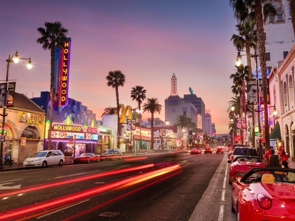 Road lined with parked sports cars and palm trees with neon signs on theatre buildings