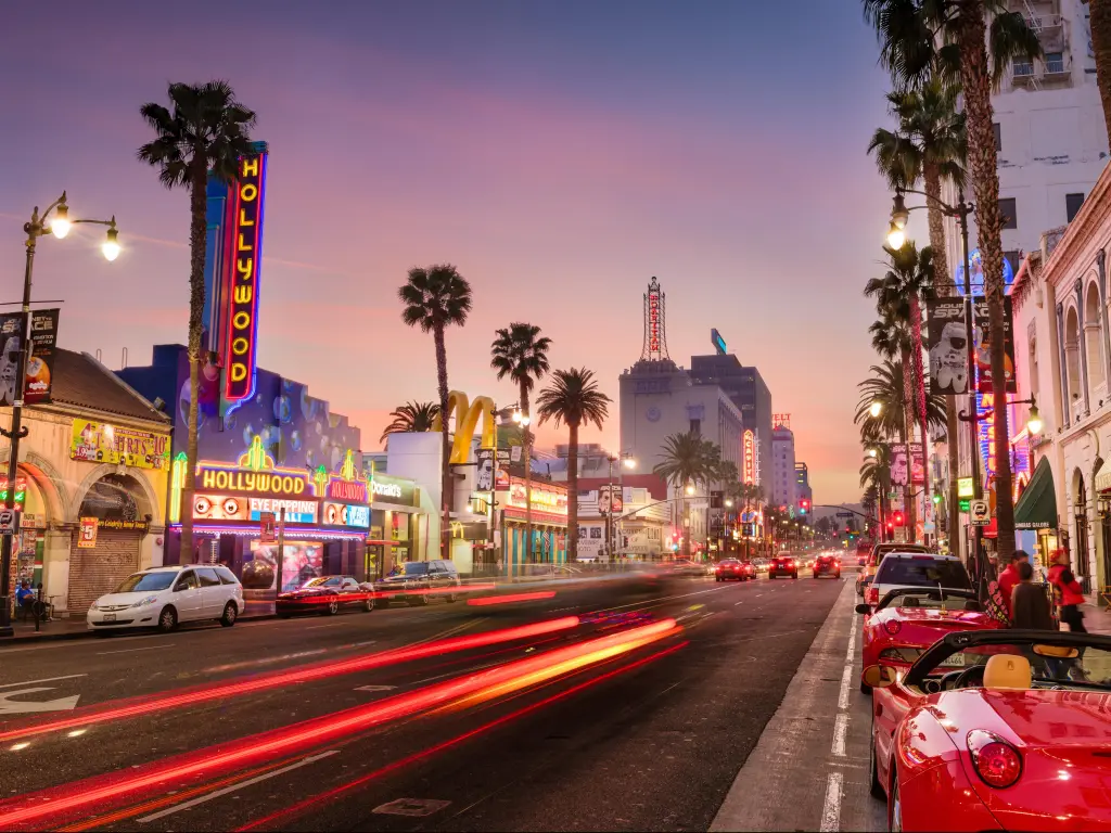 Road lined with parked sports cars and palm trees with neon signs on theatre buildings