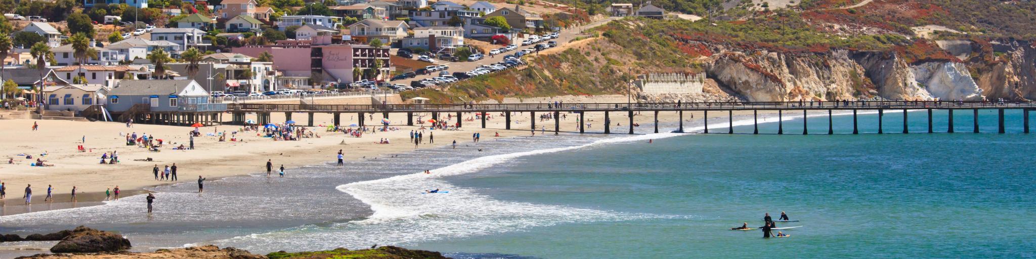 View across the ocean at Avila Beach, with the sandy shores and coastline dotted with sunbathers and swimmers, and pier jutting into the water