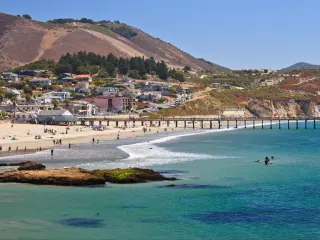 View across the ocean at Avila Beach, with the sandy shores and coastline dotted with sunbathers and swimmers, and pier jutting into the water