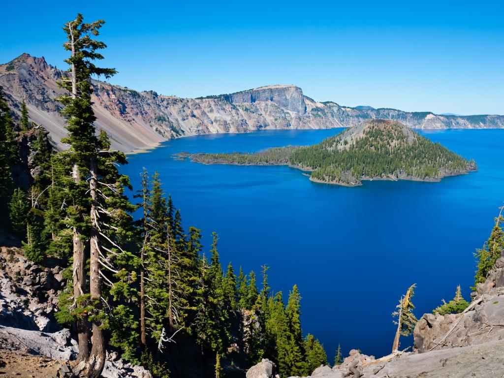 Crater Lake National Park, Oregon, USA taken on a sunny day with rocks in the foreground and the lake and mountains in the distance with a blue sky above.