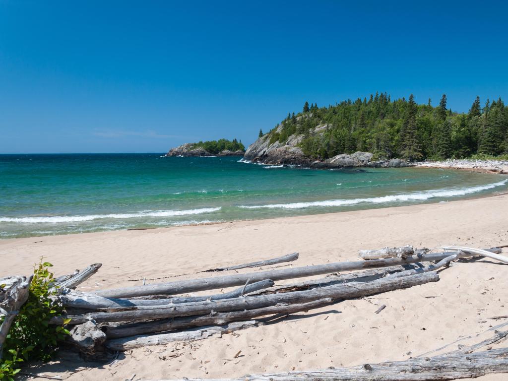 Pukaskwa National Park of Canada, Ontario, Canada taken at the northern shore of Great Lake Superior with driftwood on the sand in the foreground, calm waters and tall trees in the distance on a sunny day.