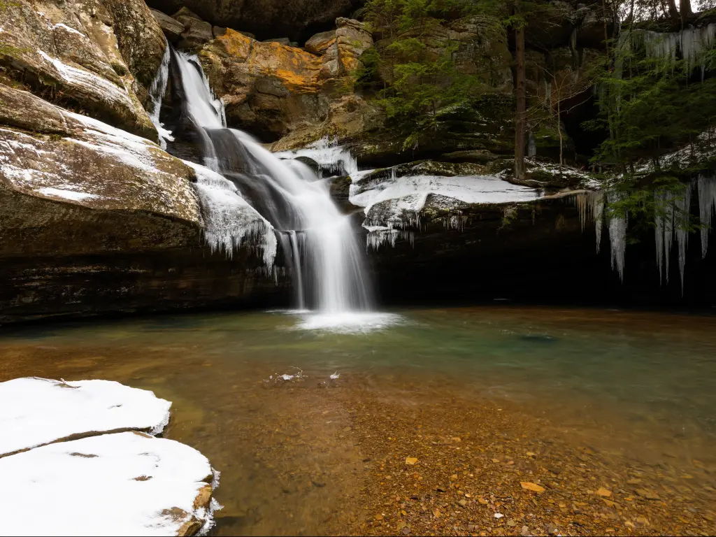 A small waterfall cascades over brown rocks into a calm pool, with large icicles forming on the rocks